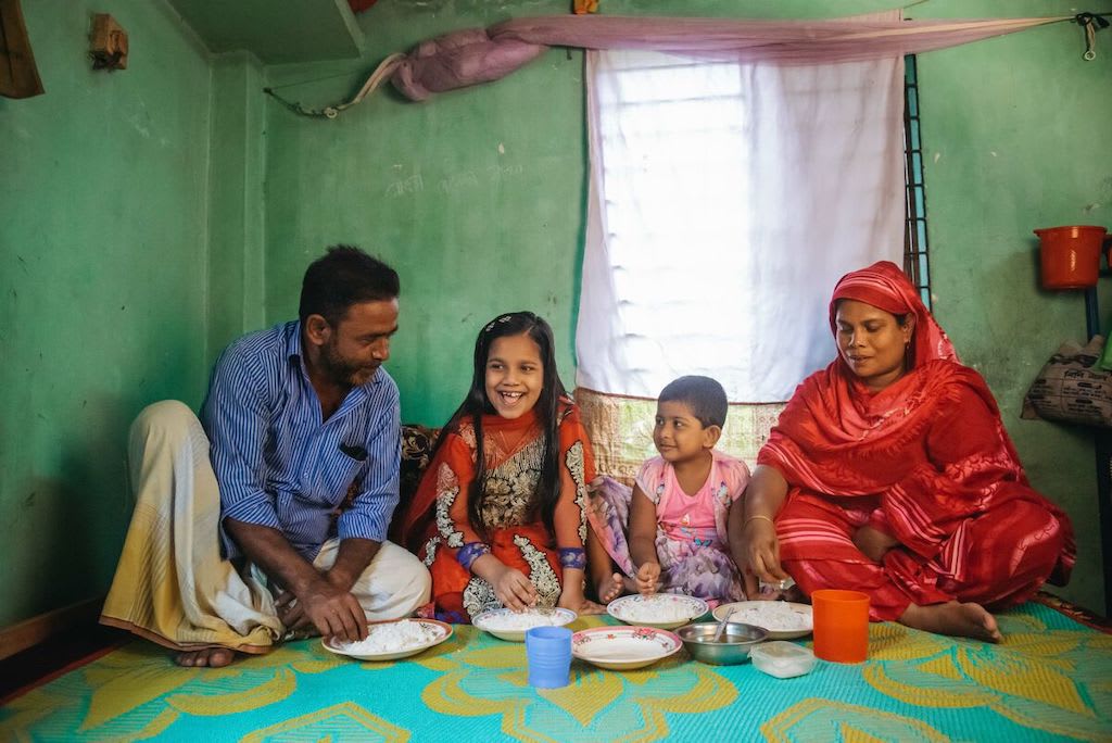 Mim is wearing an orange and purple dress. She is with her mother, wearing red, father, wearing a blue shirt and yellow pants, and her younger sister, wearing pink. They are sitting on the floor of their home and are eating a meal together. The wall behind them is green.